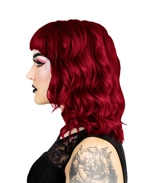 Denne profil viser en person med knaldrødt, bølget hår ved hjælp af Hermans Hårfarve Scarlett Rouge. Fed pink og sort øjenmakeup forbedrer deres udseende, parret med sort læbestift og en synlig armtatovering. De er klædt i en sort, tekstureret ærmeløs top mod en almindelig hvid baggrund.