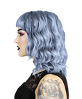 Load image into Gallery viewer, En person med lyseblåt krøllet hår og pandehår, opnået ved hjælp af Hermans Hårfarve Stella Blue (115 ml), bærer dramatisk makeup med mørk læbestift og øjenskygge. De er i en sort top med blondedetaljer og har en rosetatovering på armen mod en almindelig hvid baggrund.
