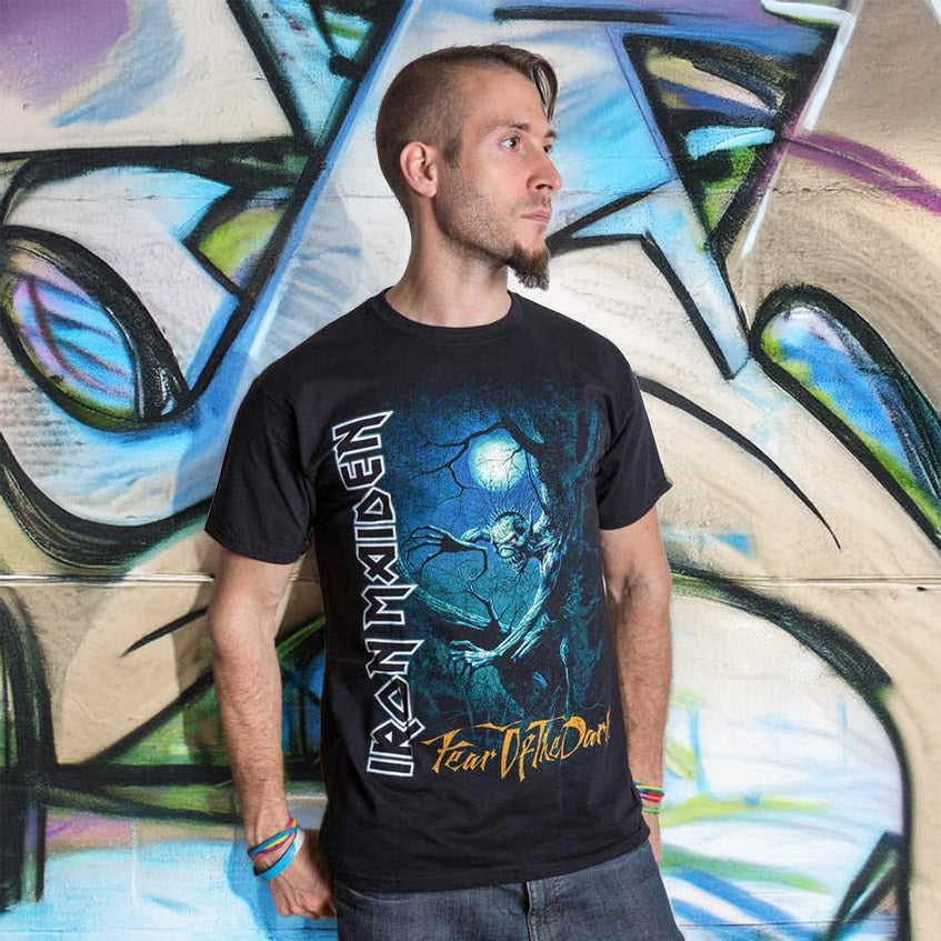 En mand med trimmet skæg og kort hår står foran graffiti-beklædte vægge. Han bærer en sort Iron Maiden "Fear of the Dark"-t-shirt og farverige armbånd og kigger til siden med hænderne i lommerne.