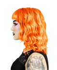 Load image into Gallery viewer, En person med lyst orange, bølget hår i bob-stil og pandehår fremviser Hermans Hårfarve Tara Tangerine (115 ml) af mærket Hermans. De bærer dramatisk makeup med sort læbestift og lyserød øjenskygge, og en tatovering er synlig på deres arm mod deres sorte blonde-detaljerede top, sat mod en hvid baggrund.

