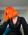 Load image into Gallery viewer, En person med Hermans Hårfarve Tara Tangerine lyst orange hår stylet i en bølget bob vender væk fra kameraet, iført en sort top og lædersele. I baggrunden er der et glimt af et køkkenområde i et hvidvægget rum.
