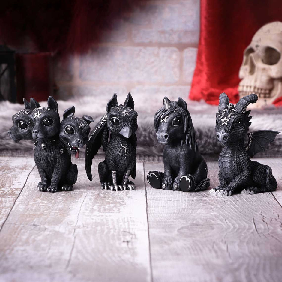Fire små dyrefigurer i gotisk stil med sorte fjer, skæl eller pels står på række på et trægulv. De har mystiske designs, såsom stjerner og horn. Et kranium og et rødt gardin er synligt i baggrunden.